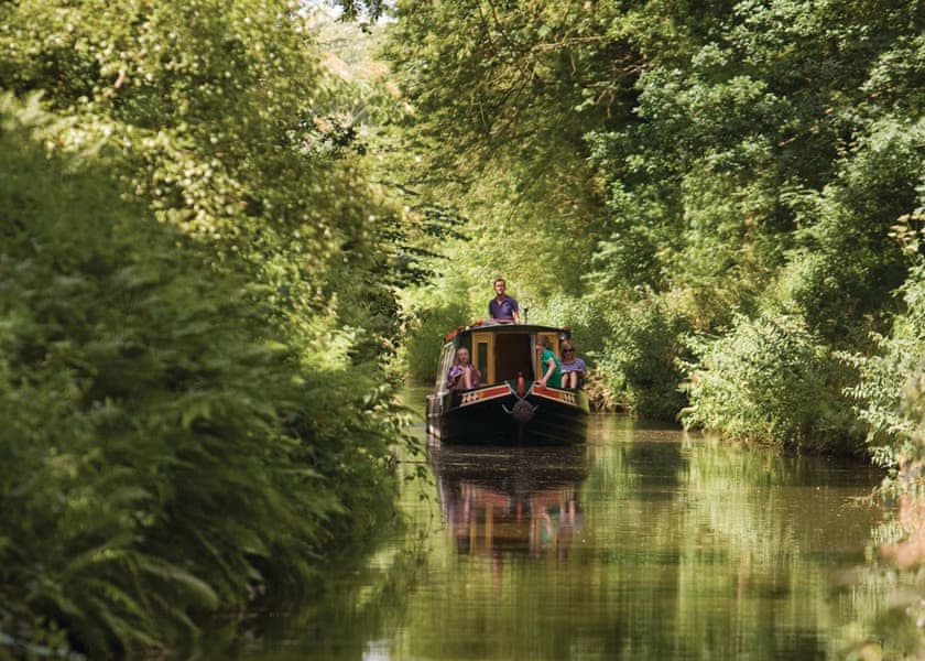 A narrowboat cruising through leafy English canal countryside.